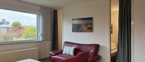 Een kamer met een rode leren bank, een houten tafel, een schilderij aan de muur en uitzicht door een raam.
