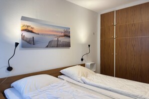 A hotel room with a bed, a painting of a beach scene, a wooden wardrobe, and a wall-mounted speaker. - Bungalow mit Garten in Dornumersiel (Dornumersiel)