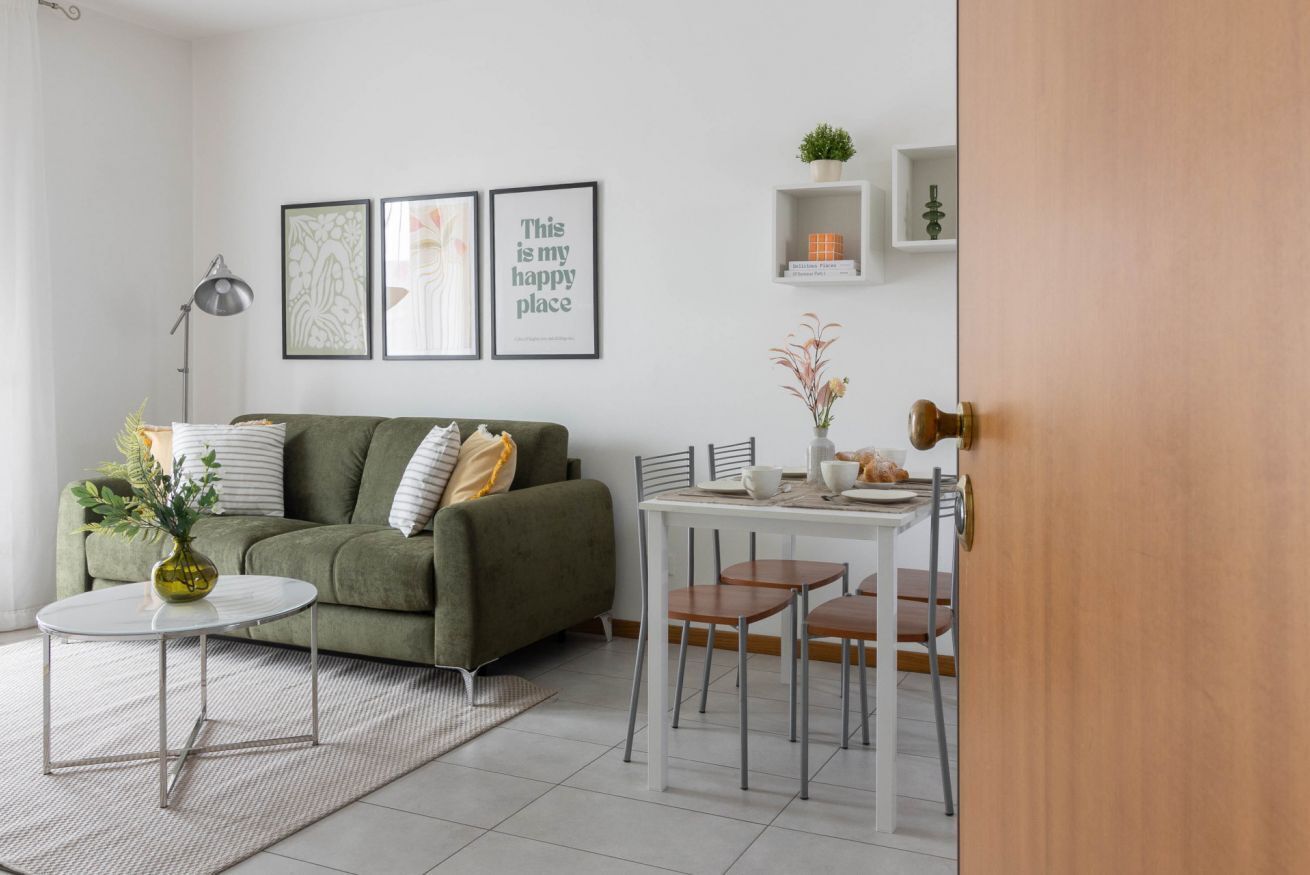A modern living room with a green sofa, a round coffee table, and a dining table with chairs. There are framed pictures on the wall and a small plant on a shelf.