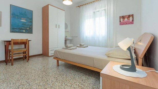 Ein Schlafzimmer mit Bett, Schreibtisch, Kleiderschrank, Nachttischlampe und einem Fenster mit Vorhängen.