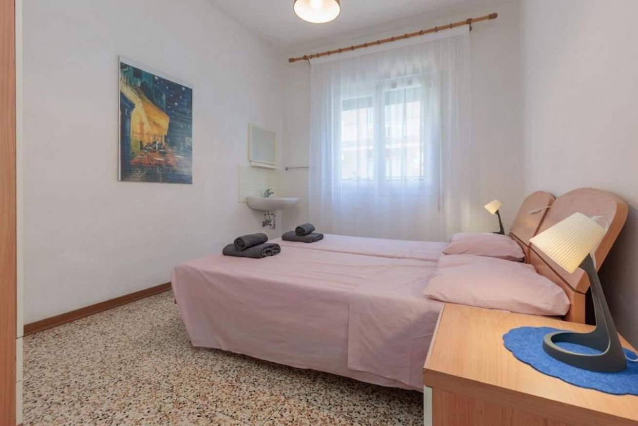 Une chambre avec deux lits, une table de chevet avec une lampe, un tableau au mur et une fenêtre avec des rideaux légers.