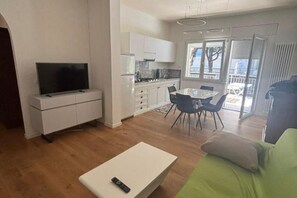 Ein modernes Wohnzimmer mit einem Flachbildfernseher auf einem weißen Schrank, einer Essecke mit Tisch und Stühlen sowie einem grünen Sofa.