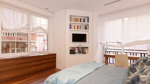 Een slaapkamer met een houten bankje, een bed met een blauwe deken, een boekenkast met boeken, een televisie en een balkon met een leuning.