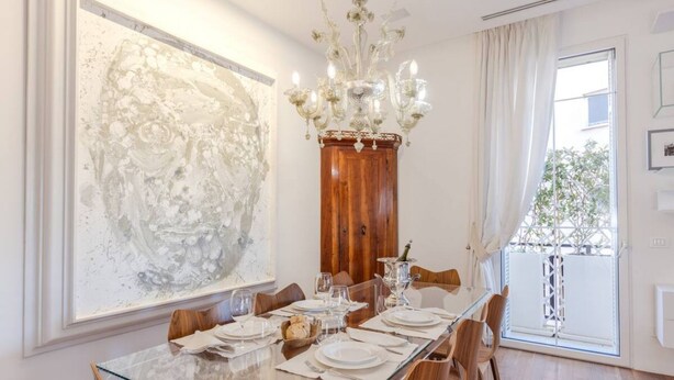 Een eetkamer met een glazen tafel, houten stoelen, een kroonluchter en een groot abstract schilderij aan de muur.