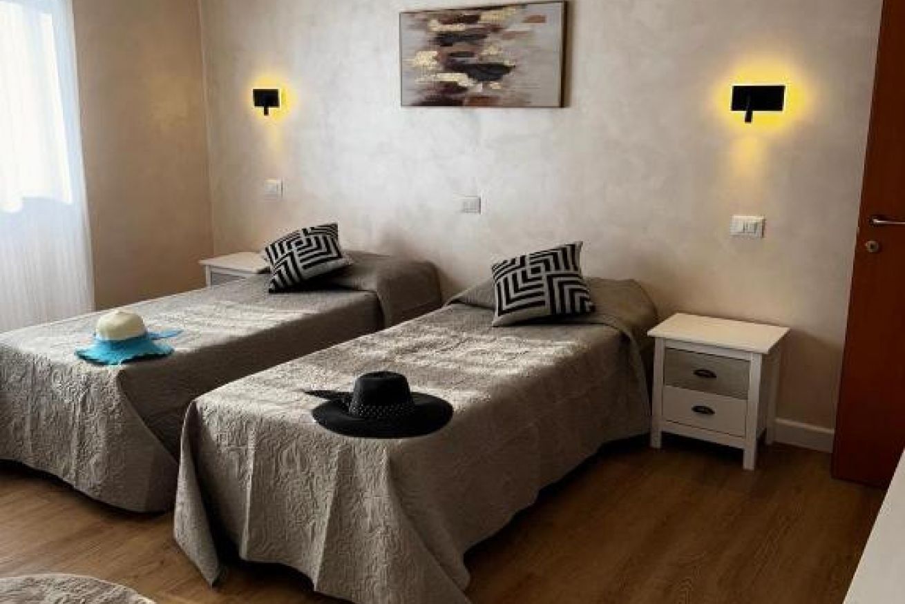 Une chambre avec deux lits, chacun étant agrémenté d’un oreiller et d’un chapeau posé dessus. On y trouve également des tables de chevet et des lampes fixées au mur.