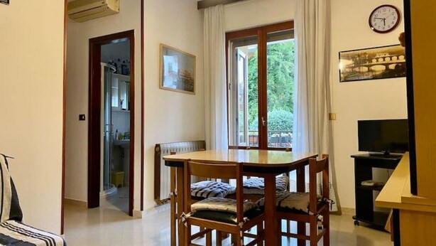 A dining area with a wooden table and chairs, a window with curtains, a clock on the wall, and a television on a stand. - Sottomarina - House Near Sea | Terrace & Private Garage (Chioggia)