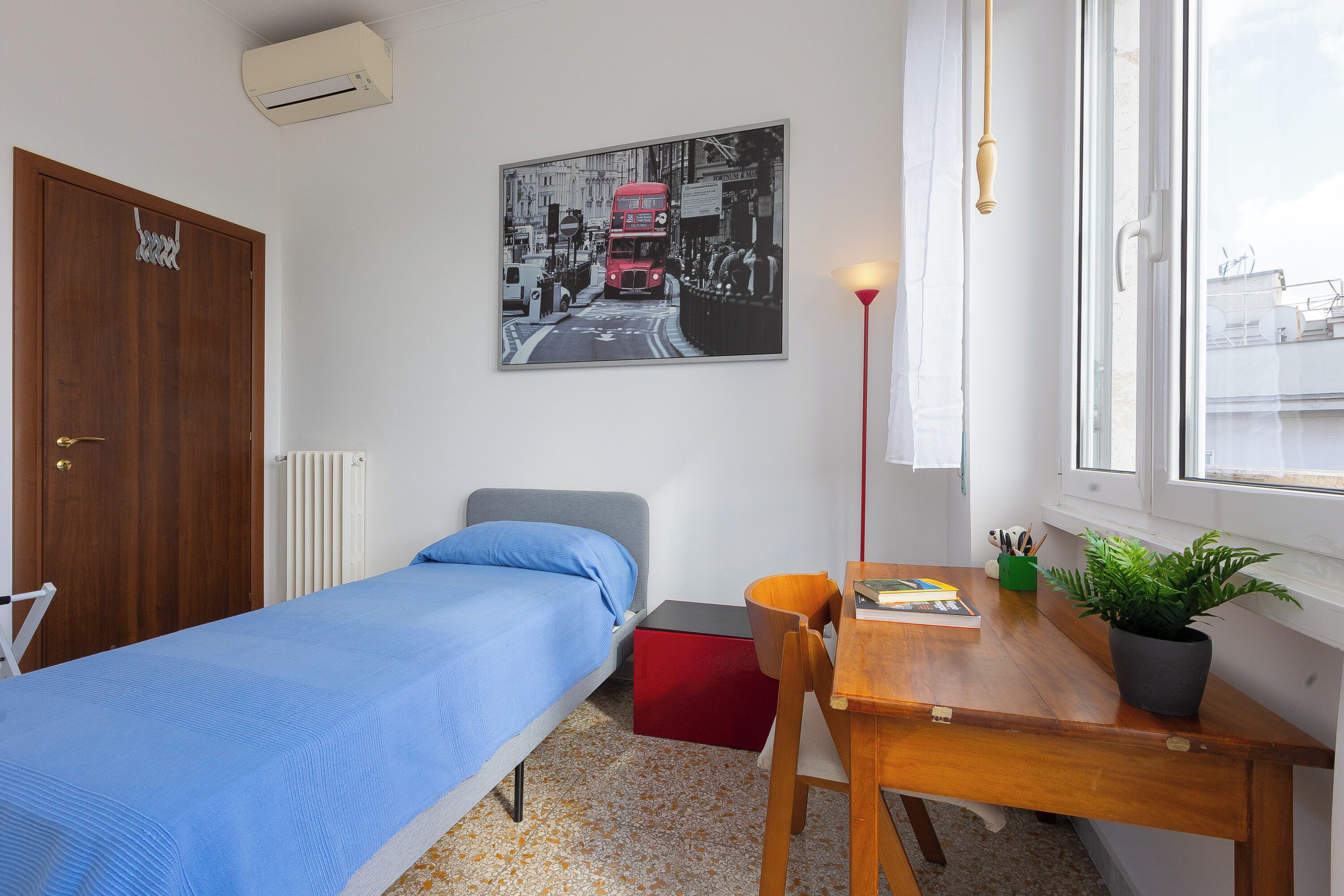 A bedroom with a single bed, a small desk, a chair, a radiator, a window with a view, and a wall-mounted air conditioner.