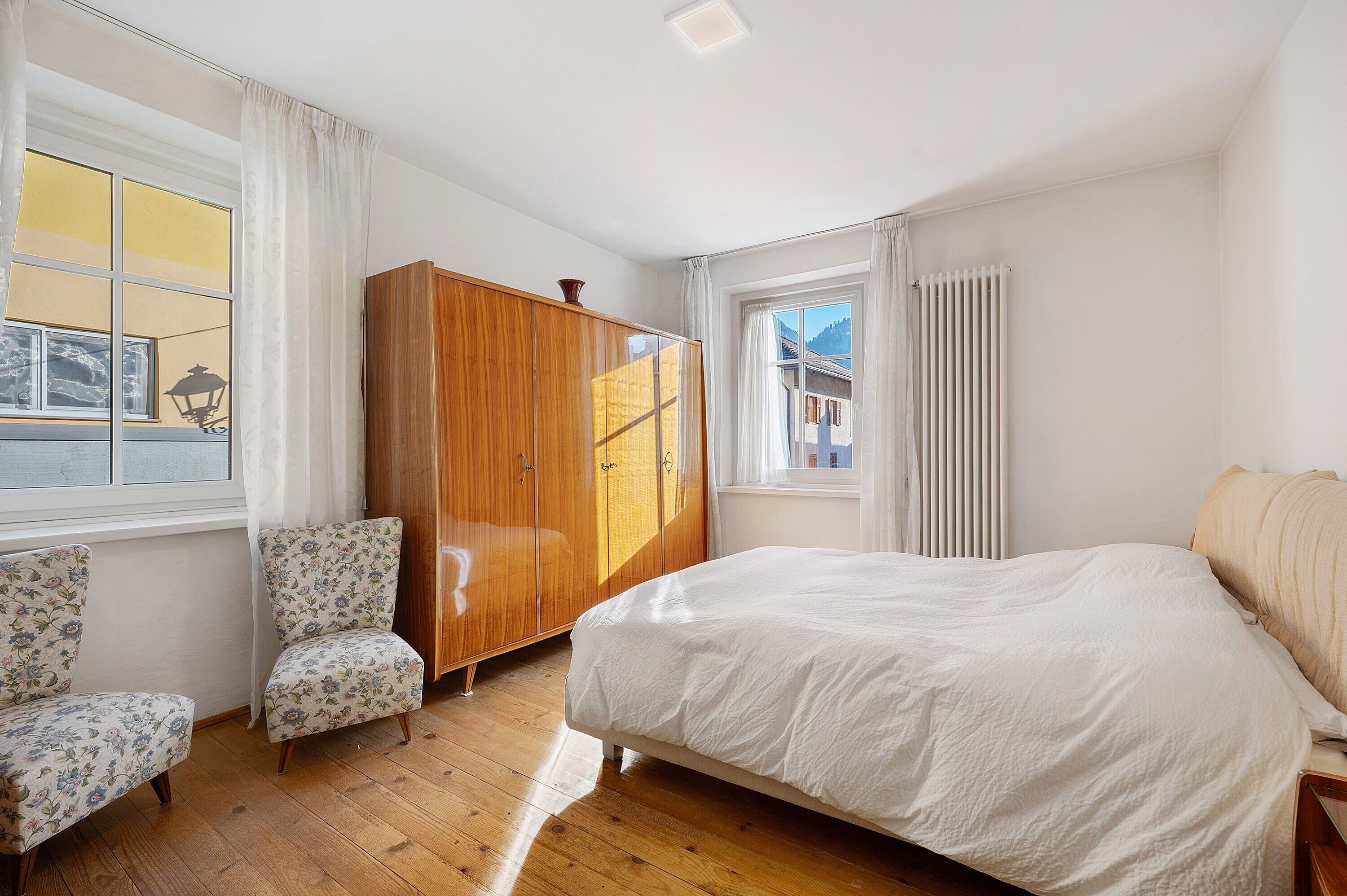 Un dormitorio con una cama, un armario de madera, dos sillones con estampado floral y una ventana con cortinas.