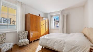 Un dormitorio con una cama, un armario de madera, dos sillones con estampado floral y una ventana con cortinas.