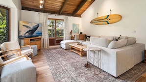 A living room with a wooden ceiling, a sofa, a coffee table, a TV, and a surfboard on the wall. - Large 3BR + Loft at Kanaloa Pools, Golf Views & Prime Keauhou Location (Kailua-Kona)