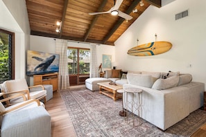 A living room with a wooden ceiling, a sofa, a coffee table, a TV, and a surfboard on the wall. - Large 3BR + Loft at Kanaloa Pools, Golf Views & Prime Keauhou Location (Kailua-Kona)