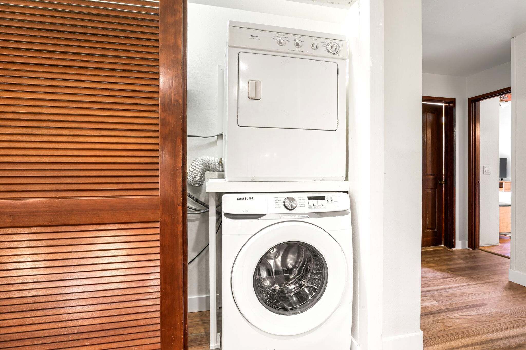 Une buanderie équipée d’un lave-linge et d’un sèche-linge Samsung, de stores en bois et d’un couloir menant aux autres pièces.
