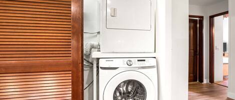 Une buanderie équipée d’un lave-linge et d’un sèche-linge Samsung, de stores en bois et d’un couloir menant aux autres pièces.