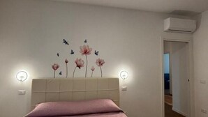Makuuhuone, jossa on sänky, seinälle asennettu ilmastointilaite ja ovi, joka johtaa toiseen huoneeseen.