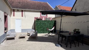 Une cour avec un toit en tuiles, une porte blanche, une haie verte, du mobilier d’extérieur et une table avec des chaises.