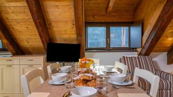 Zona pranzo con soffitto in legno, televisore e tavolo apparecchiato con ciotole, bicchieri e pasticcini.