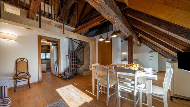 Een ruime loft met hoge plafonds, zichtbare houten balken, een eethoek met tafel en stoelen, een keuken en een trap naar een hoger niveau.