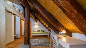 Slaapkamer in een loft met zichtbare houten balken, een wit bureau, een stoel, een bed met groene kussens en een nachtkastje met een lamp.