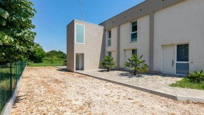 Un edificio moderno con techo plano, una pequeña estructura cuadrada con ventana y un área pavimentada con grava y plantas pequeñas.