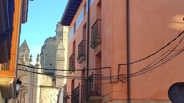 A narrow street with historic buildings, including a stone structure with a pointed roof and a building with a flat roof and balconies. - Casa palaciega Doña Leocadia (Viana)
