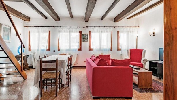 Ein geräumiges Wohnzimmer mit einem roten Sofa, Holzbalken an der Decke, einem Esstisch mit Stühlen und einer Treppe.