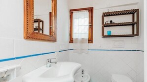 Ein Badezimmer mit einem weißen Waschbecken, einem Bidet und einer Toilette. Darüber hängt ein Spiegel mit Holzrahmen, und darunter befindet sich ein Ablagefach mit einem Föhn.