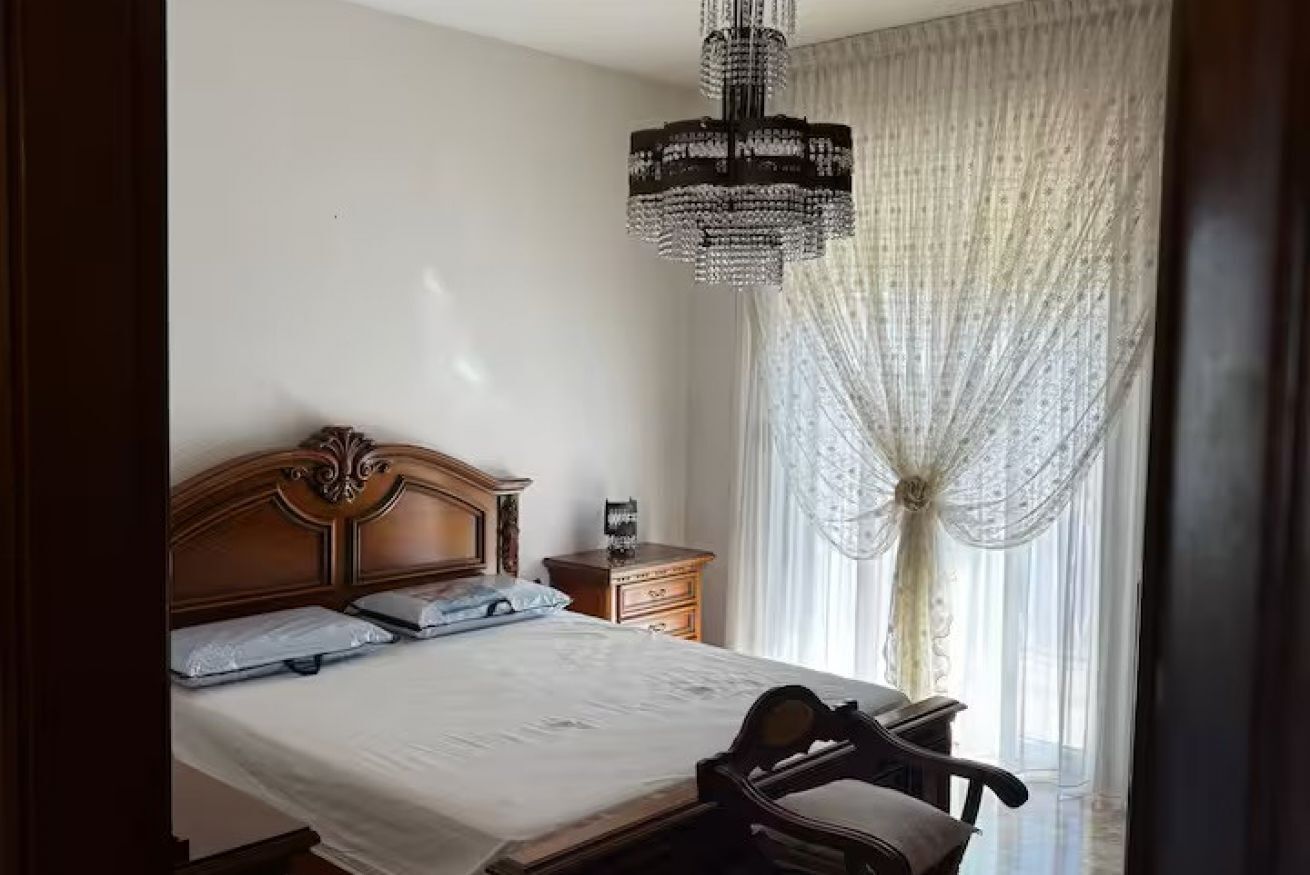 Quarto com cabeceira de madeira, mesa de cabeceira, cadeira, lustre e cortinas translúcidas.