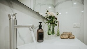 Casa de banho com lavatório de mármore branco, uma torneira moderna, dispensador de sabonete e um vaso com flores.