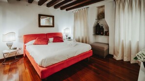 Quarto com cama vermelha, roupa de cama branca, piso de madeira, uma pequena mesa de cabeceira com abajur, um espelho na parede e uma estante com objetos decorativos.