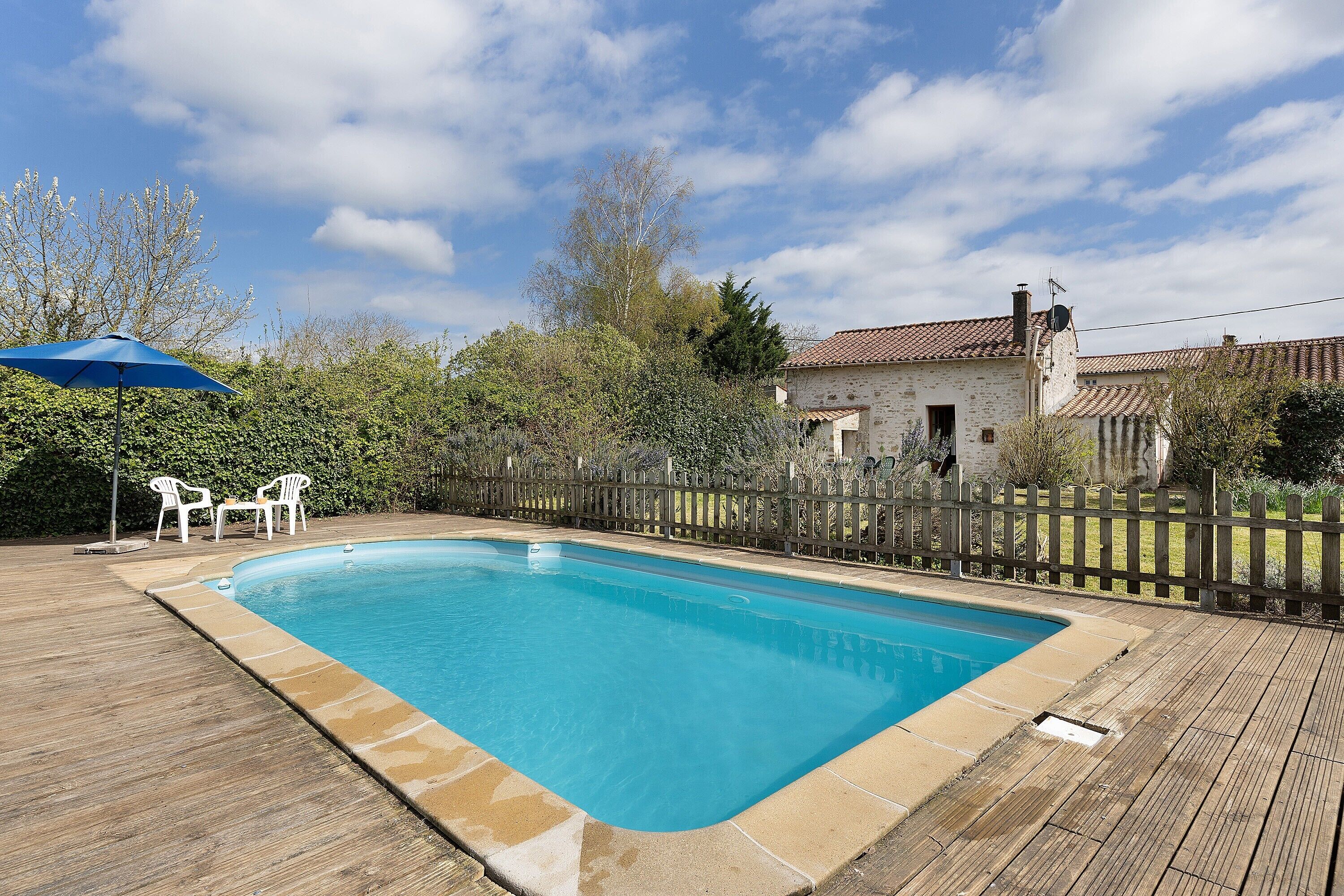 A rectangular swimming pool with a wooden deck, surrounded by a wooden fence, and a white building with a red-tiled roof in the background.