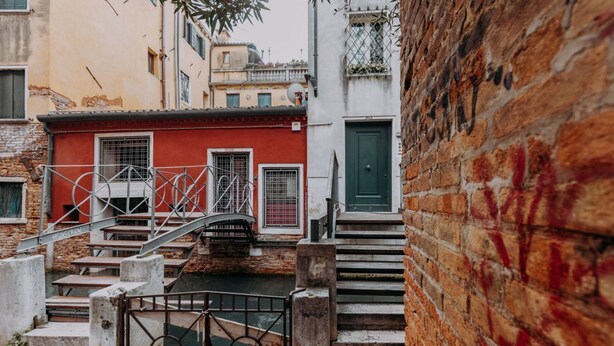 Un callejón estrecho con un edificio rojo, una puerta verde y una escalera metálica que lleva a un puente sobre un canal.