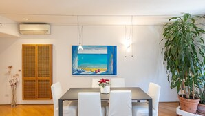 A dining area with a table, chairs, a painting, an air conditioner, and a potted plant. - Rental in Rome San Gioacchino View Apartment (Rome)