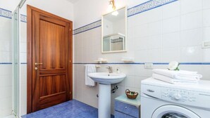 Een badkamer met een houten deur, een wastafel op voet, een spiegel, een wasmachine en een klein tafeltje met een mandje.