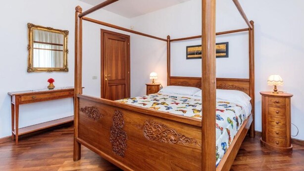 Een hemelbed met een bloemenbedekking, een houten nachtkastje met een lamp, een houten dressoir en een spiegel aan de muur.