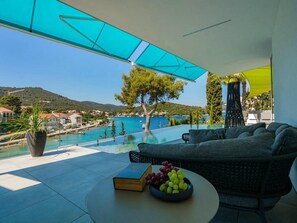 A terrace with a view of a coastal town, an infinity pool, and a seating area with a book and grapes.