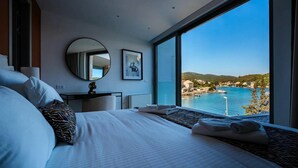 Habitación de hotel moderna con una cama grande, un escritorio, una silla, un espejo redondo y vistas a un pueblo costero y al mar.