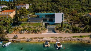 Una casa moderna con un amplio balcón que da a un mar azul y despejado, rodeada de exuberante vegetación y otros edificios.