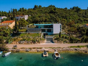 A modern house with a large balcony overlooking a clear blue sea, surrounded by lush greenery and other buildings.