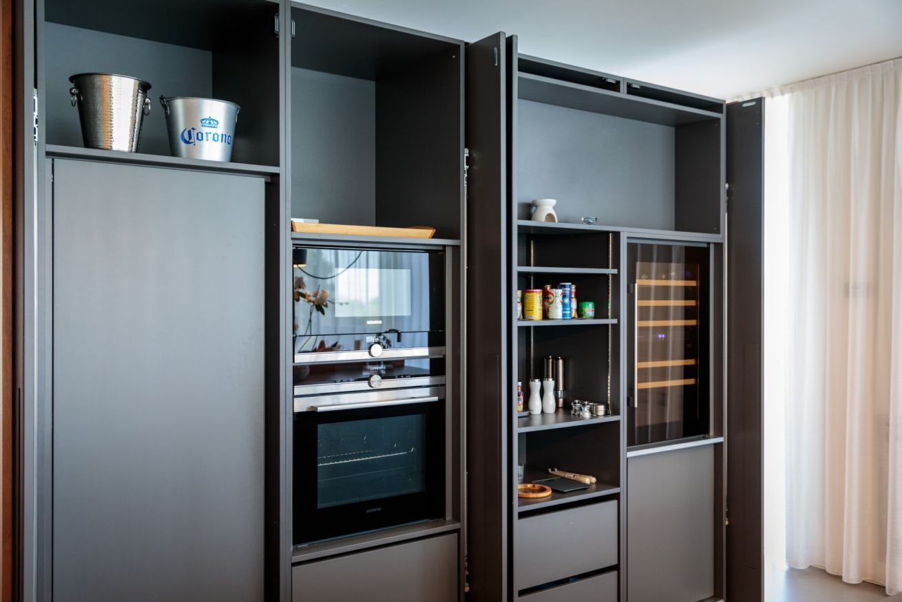 A modern kitchen with a built-in oven, a microwave, and a coffee machine. There are shelves with various kitchen items and a cabinet with a silver bucket and a can of beer.