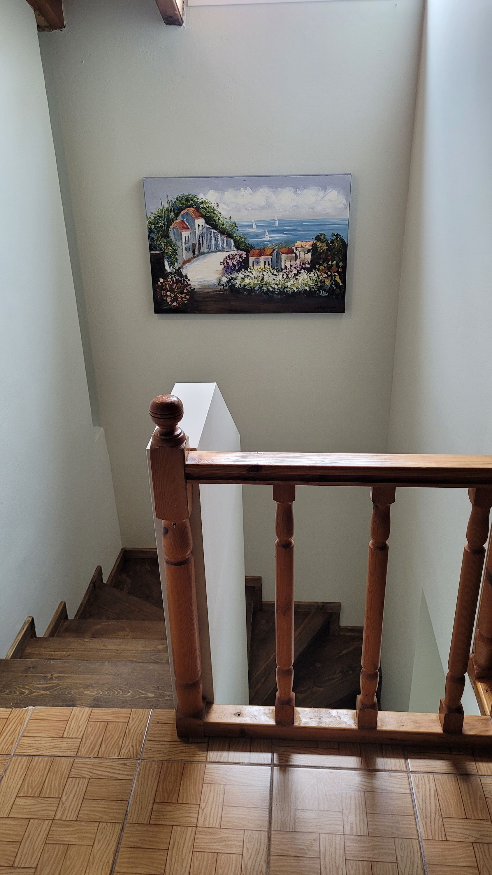 A wooden staircase with a railing, a painting of a coastal scene on the wall, and a herringbone-patterned floor.