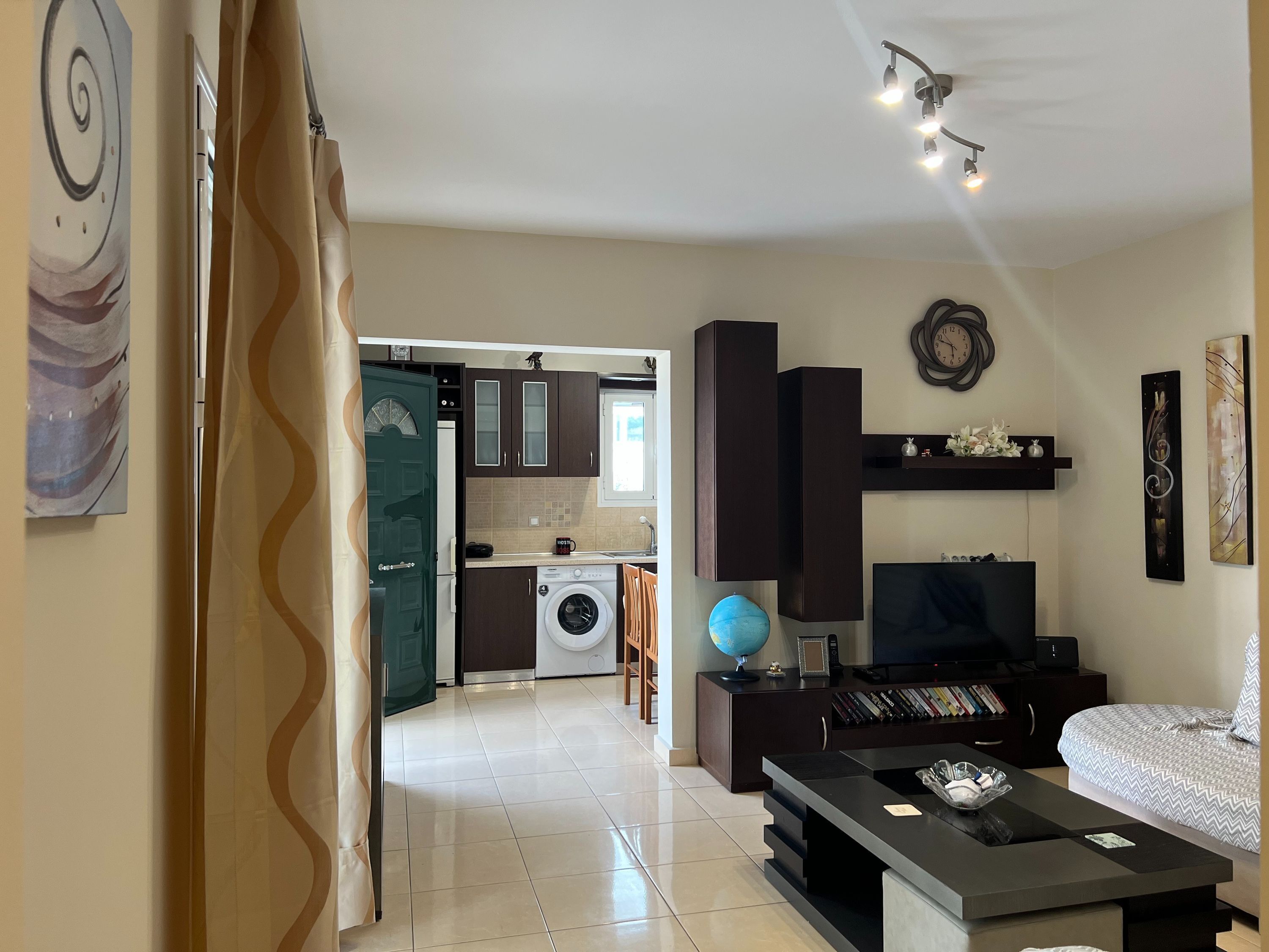 A modern living room with a sofa, a coffee table, and a TV. There is a kitchen in the background with a washing machine and a dining area.