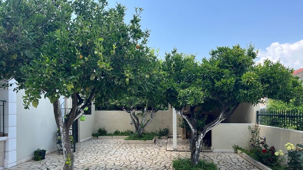 A courtyard with stone flooring, trees, and a white wall. - Kika Residence - 2 Bedroom House With Barbeque (Kefalonia)