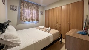 A bedroom with a bed, a wooden wardrobe, a desk, and a window with floral curtains. - Kika Residence - 2 Bedroom House With Barbeque (Kefalonia)