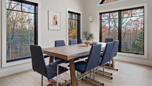 Uma sala de jantar com uma mesa de madeira e cadeiras azuis, grandes janelas com vista para a folhagem outonal e um quadro na parede.
