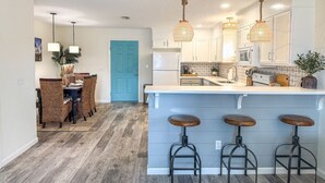 A modern kitchen with a central island, bar stools, pendant lights, and a dining area with wicker chairs. - 8th At The Bay, New Listing (Port St. Joe)