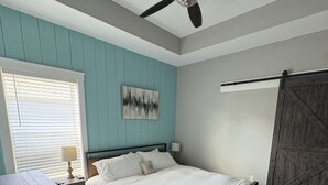 Un dormitorio con cama, ventilador de techo, ventana con persianas, una obra de arte en la pared y una puerta corredera estilo granero.