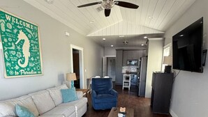 Una sala de estar con ventilador de techo, un sofá, un sillón azul, una mesa de centro y una cocina al fondo.