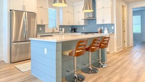Eine moderne Küche mit weißen Schränken, einem Edelstahl-Kühlschrank, einer zentralen Kochinsel mit Barhocker und Pendelleuchten.