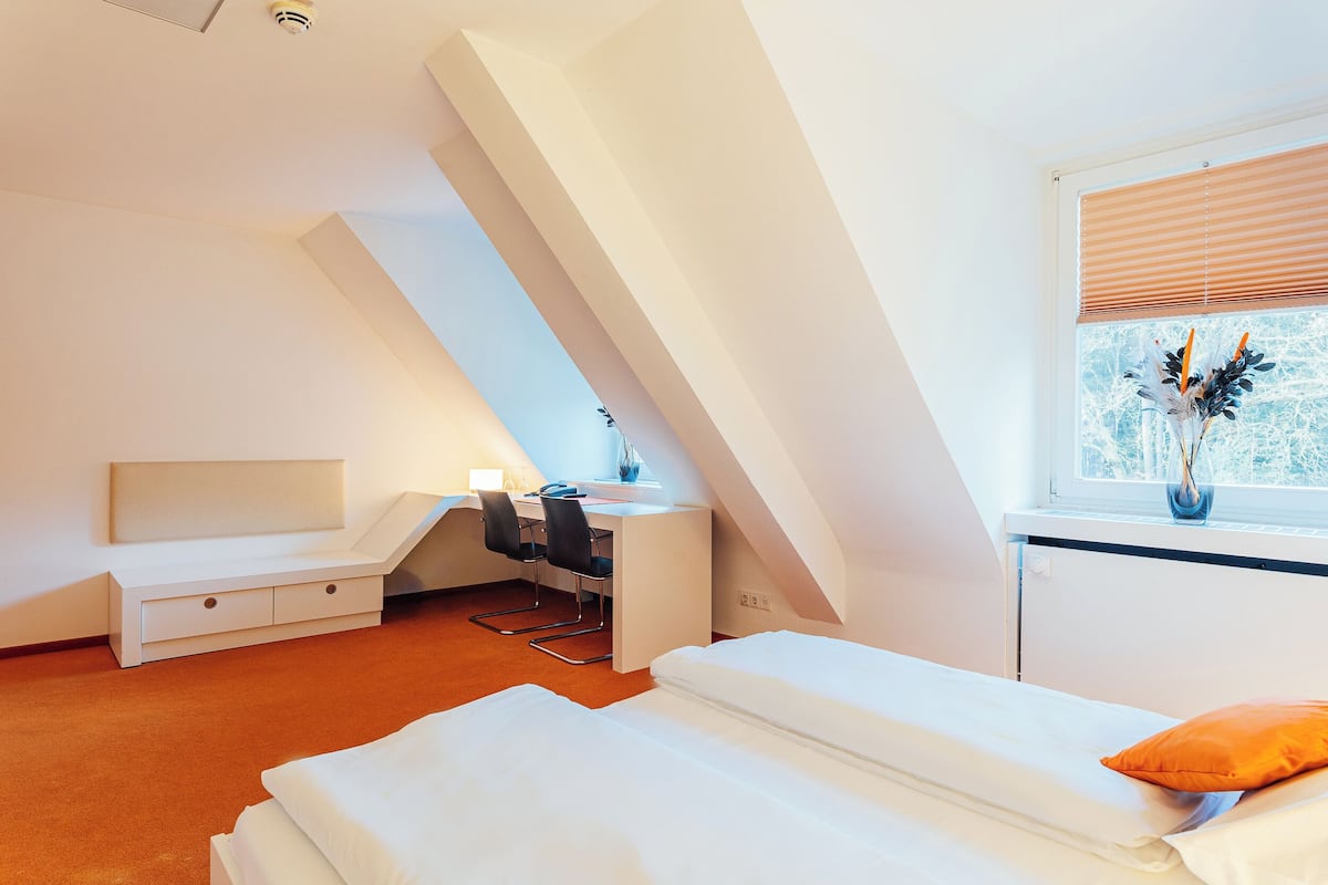 Ein modernes, helles Zimmer mit Schrägen, einem Bett mit weißer Bettwäsche, einem Schreibtisch mit Stuhl und einem Fenster mit Jalousien.