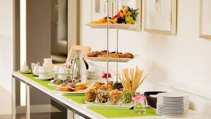 Ein reichhaltiges Buffet mit verschiedenen Speisen und Geschirr, darunter Früchte, Gebäck und Soßen, vor einer weißen Wand mit gerahmten Kunstwerken.